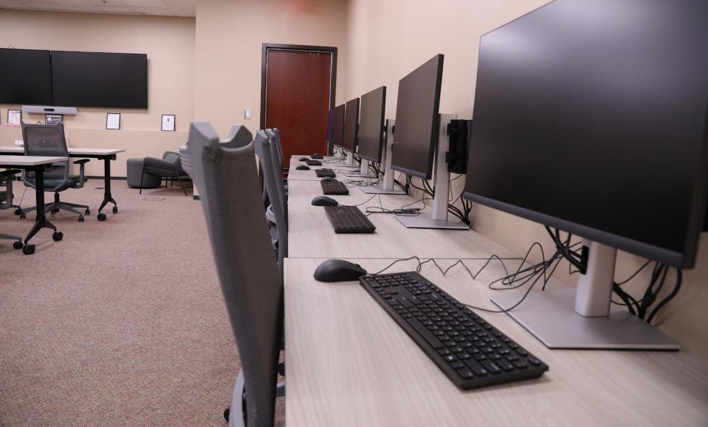 A picture showing the length of a table holding four computers against a wall. Two flat screen tvs are on the back wall with rows of tables and chairs in the center of the room.