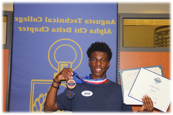 非裔美国男性, 普林斯顿·巴恩斯微笑着, 他右手拿着证书，左手拿着奖牌，站在一张蓝紫色的海报前，海报上用金色字体写着“买世界杯app推荐Alpha Chi Delta分会”. 
