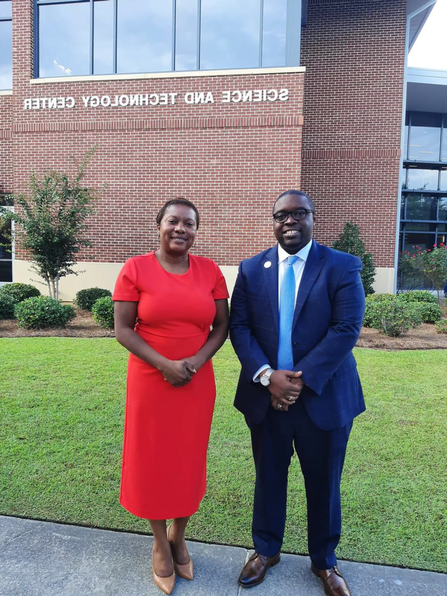 Dr. Jermaine Whirl, an African American male smiles wearing black rimmed glasses, a navy suit with a light blue tie and white shirt stands beside Kimberly Ballard-Washington, J.D., an African American Female, who is also smiling wearing a red dress. The two stand in front of the Science and Technology Center
