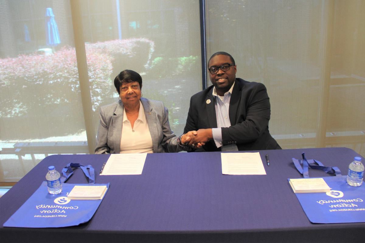 African American male wearing a blue blazer, 白领衬衫, 银色领章, sitting in a chair shaking hands next to an African American female wearing a gray blazer, 白衬衫, both are sitting in front of a table with a blue table cloth, on the table are white papers and blue and white bags. The photo is in front of a window, and outside the window are flowers.
