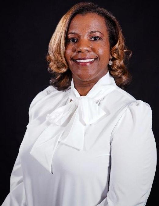 An African American female, Dr. Kristie Searcy, Dean of Health Sciences, 有一头中等长度的金色卷发，穿着白色的蝴蝶结领衬衫，戴着黑色背景的珍珠耳环，微笑着.