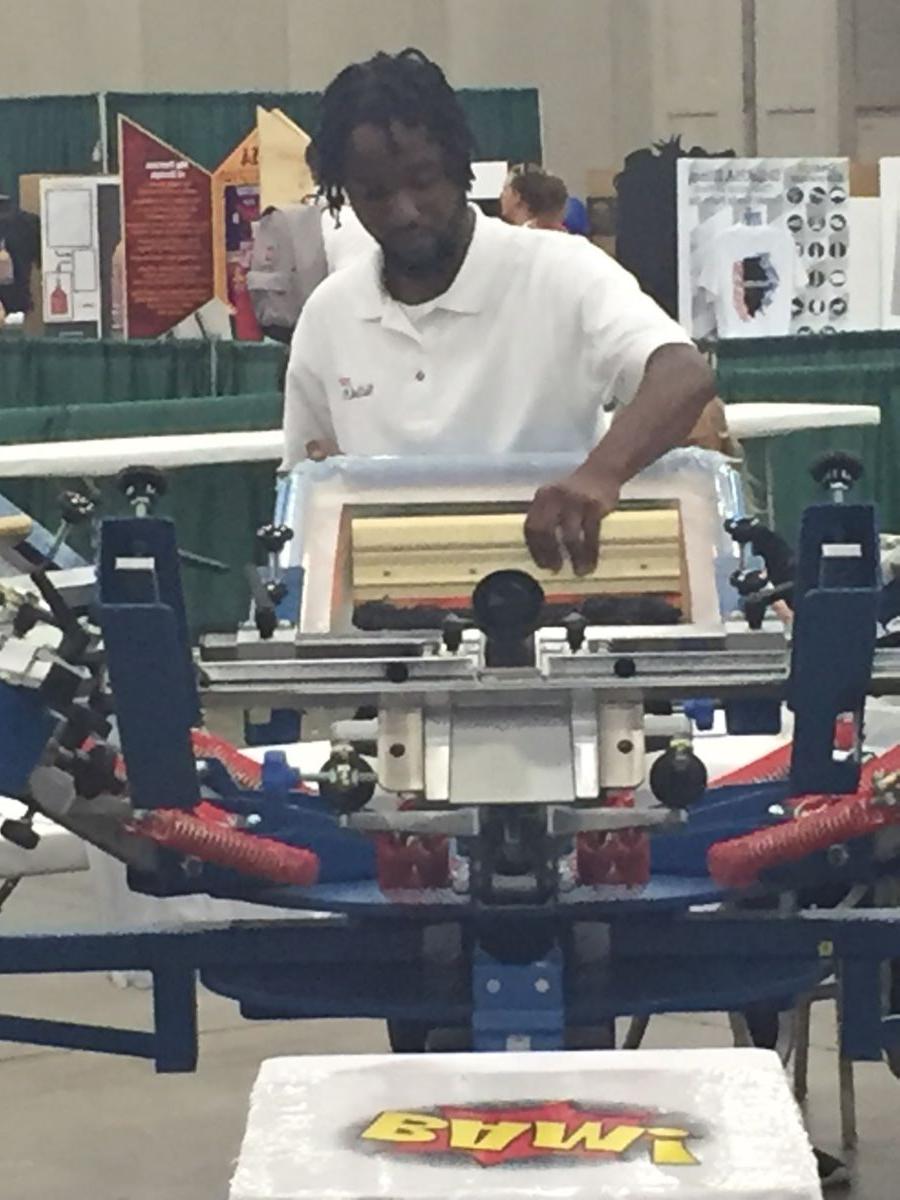 穿着白色polo衫的年轻非裔美国大学生, James McRae, 比赛期间从事丝网印刷工作.